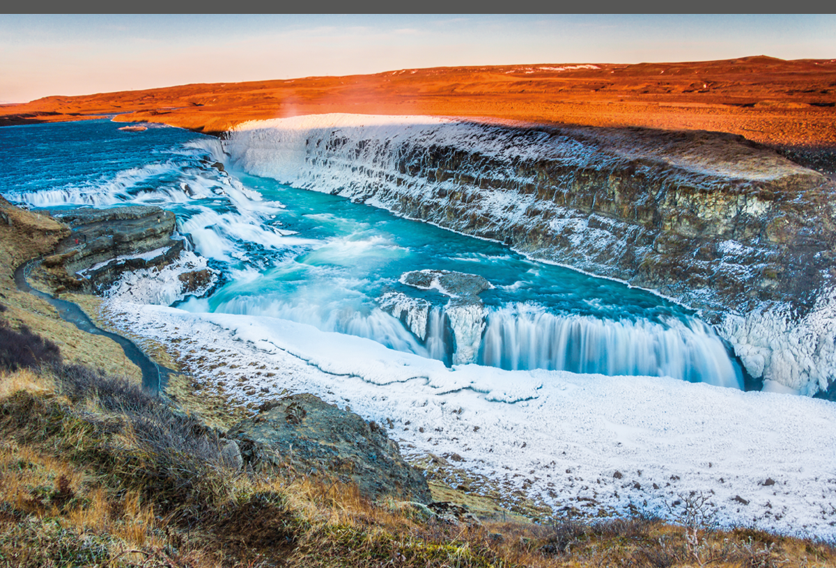 Beautiful Nordic landscape from spectacular Iceland including mighty Gullfoss waterfall partly frozen in ice as foreground and magnificent distant plane sunlit by the setting Sun for background  Very famous destination for travellers and adventurers from all over the world, this Polar country is a popular spot for tourists willing to witness and enjoy one of the wildest places in Europe  Shot with Canon EOS 60D, wide 17mm angle lens, f13, ISO 100 
