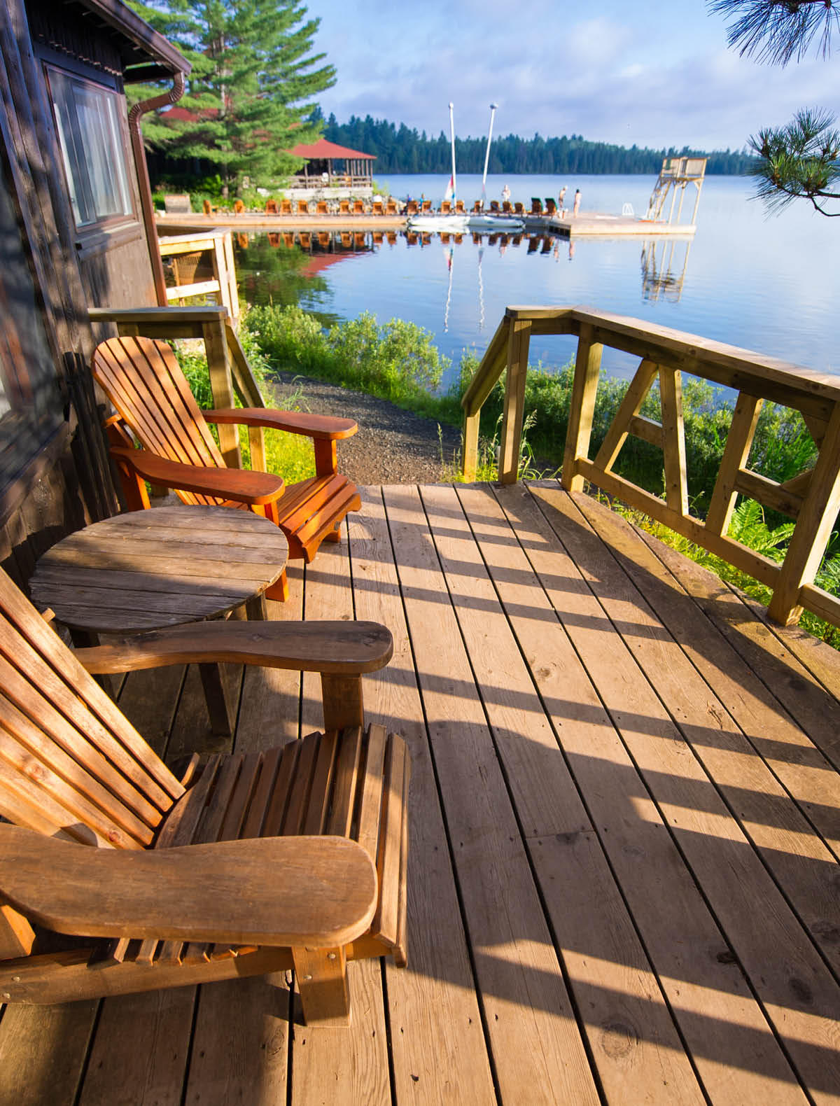 Muskoka chairs sitting on a wooden porch facing a calm lake. In the background there are Adirondack and Muskoka chairs on a wooden dock