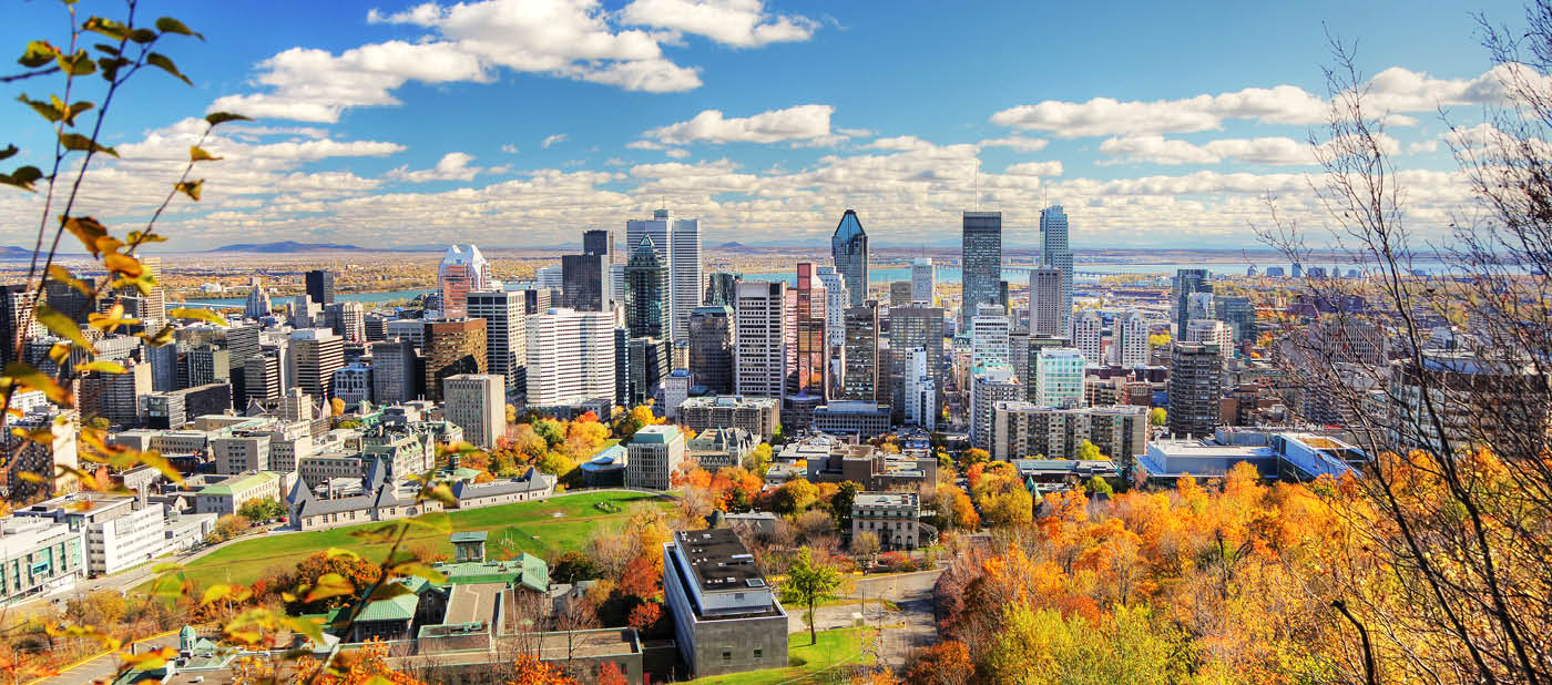 A Mountain view of Montreal City, Canada in the fall. The sky is clear and it is a sunny day.  A cluster of gray, multiple-sized buildings are visible in the center of the photo.  Lush, fall foliage lines the front of the image, and a body of water is visible in the back of the photo.