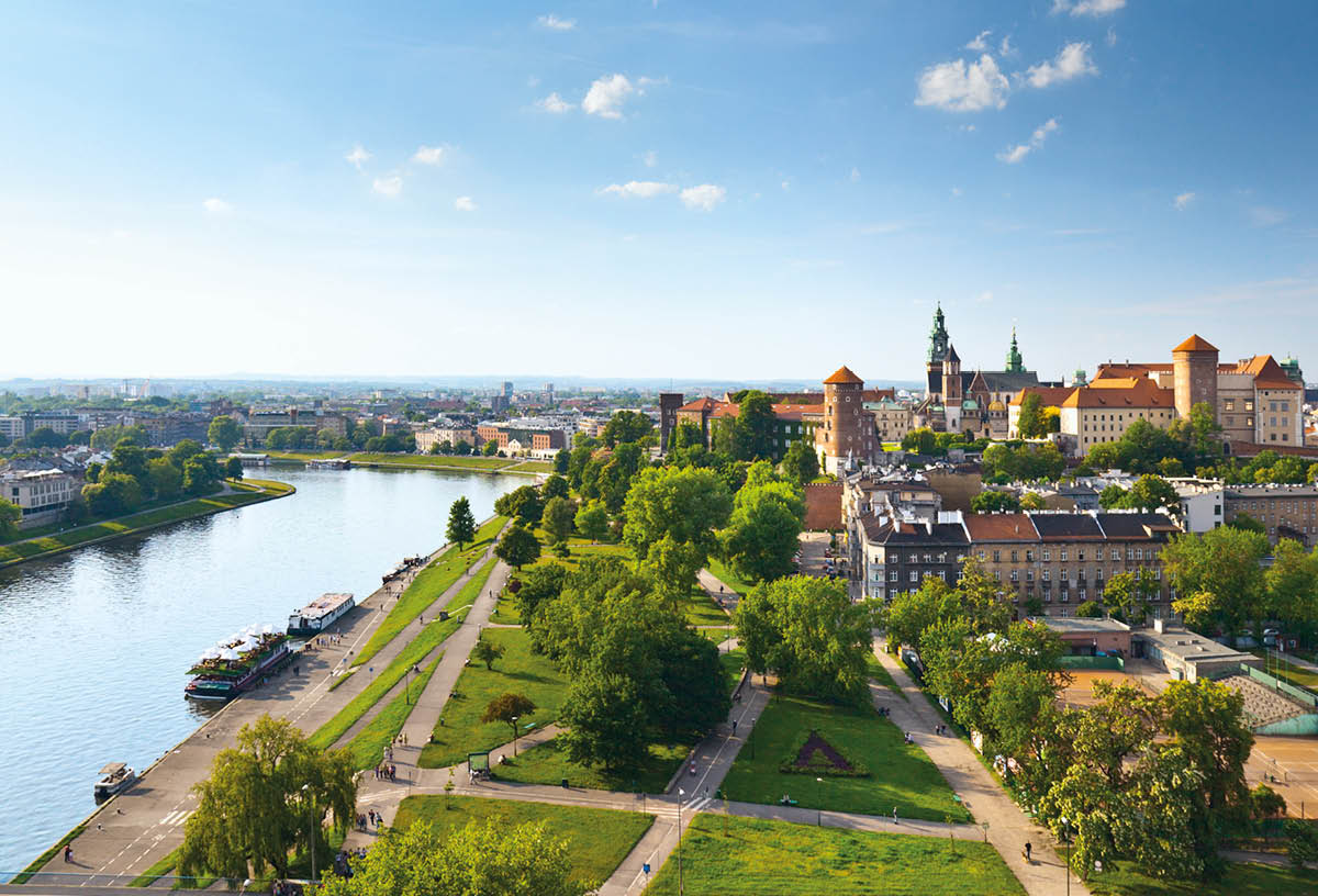 Daytime panorama of Krakow, Poland, featuring Wawel Castle   The Vistula River flows along the left side of the image, and the castle is visible on the right, near the horizon   There is a blue sky with sparse, wispy clouds above the horizon and a manicured lawn intersected by walking paths in the foreground 