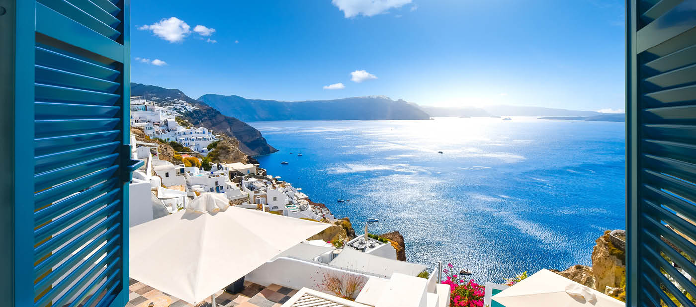 View from an open window with blue shutters of the Aegean sea, caldera, coastline and whitewashed town of Oia, Santorini, Greece.
